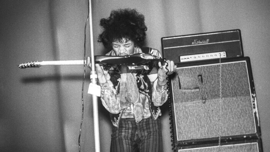 Black and white photograph of Jimi Hendrix standing by a stack of speakers and playing a guitar with his teeth.