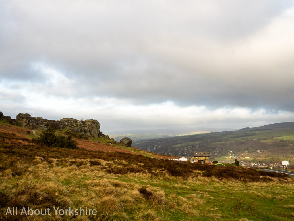 Open moorland in the foreground leading to a sandstone large rock  formation in the midground called the Cow and Calf Rocks. To the right in the background, the town of Ilkley lies in the valley.