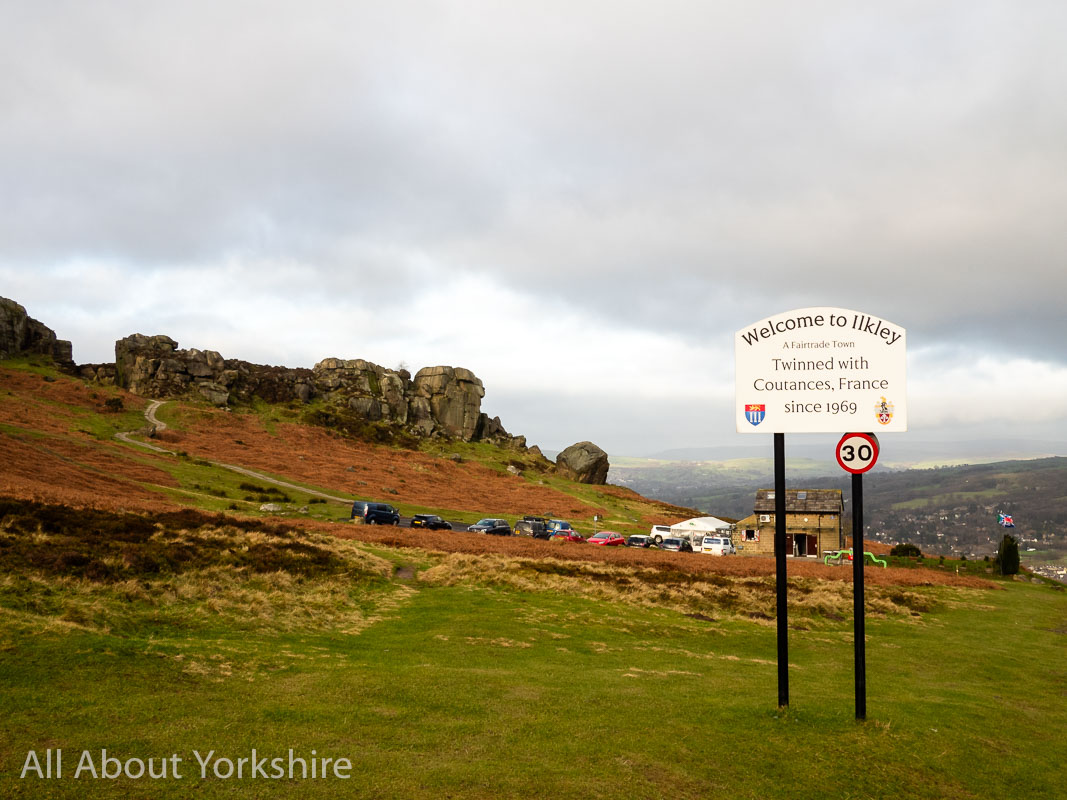 View across moorland to Cow and Calf Rocks with Welcome to Ilkley sign in foreground.