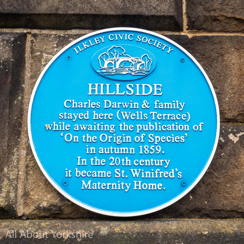 Blue plaque on stone wall. Plaque reads Ilkley Civic Society. Hillside. Charles Darwin & family stayed here (Wells Terrace) while awaiting the publication of 'On the Origin of Species' in autumn 1859. In the 20th century it became St. Winifred's Maternity Home.
