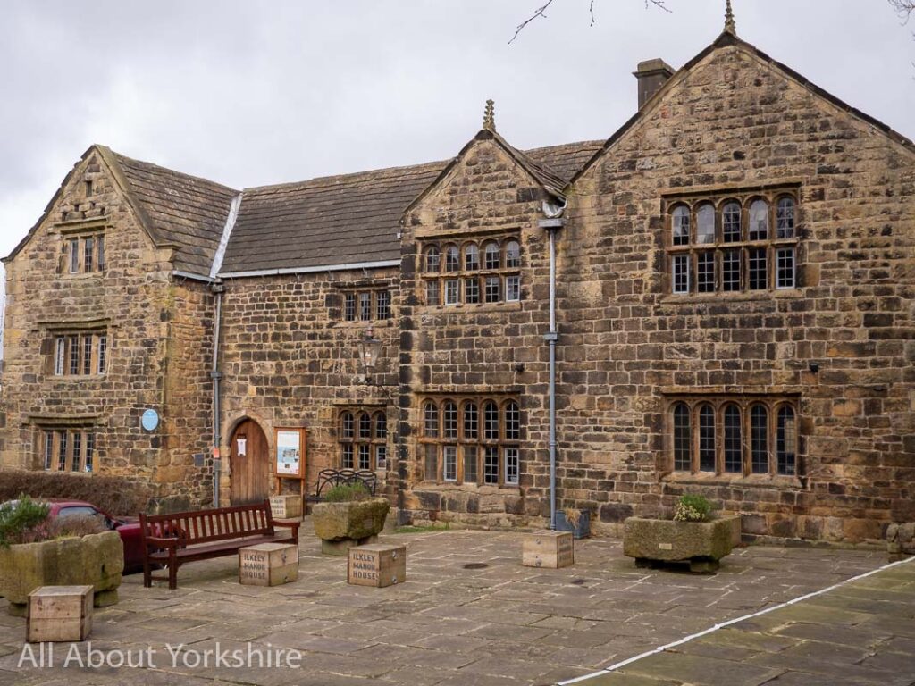 Ilkley Manor House - an old stone built manorial building with mullioned windows and a flagged courtyard and stone planters to the front.