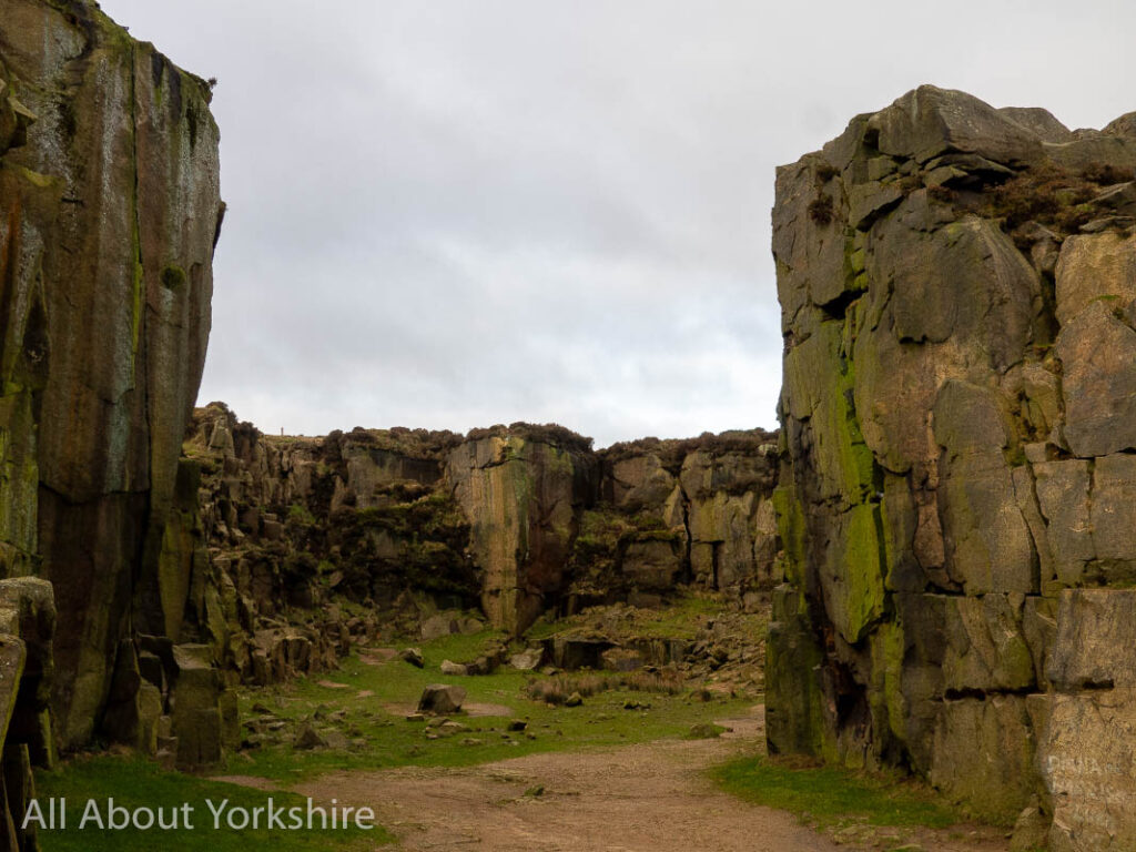 Two high stone rack faces flank a wide sandy path at the entrance to a disused quarry. 