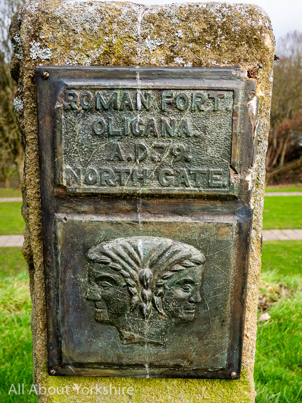 Brass plaque on small stone block that reads "Roman fort Olicana A.D.79. North Gate" Underneath the text is a relief of a two faced woman.