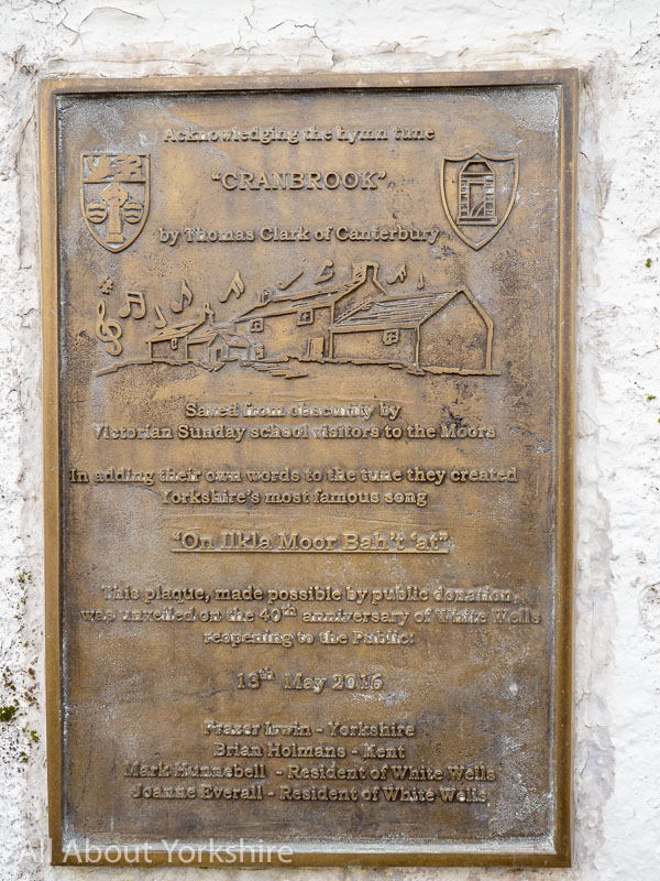 Brass plaque that states: Acknowledging the hymn tune “Cranbrook” by Thomas Clark of Canterbury. Saved from obscurity by Victorian Sunday school visitors to the Moors. In adding their own words to the tune they created Yorkshire’s most famous song “On Ilkla Moor Bah’t ‘at” This plaque, made possible by public donation was unveiled on the 40th anniversary of White Wells reopening to the Public 13th May 2016