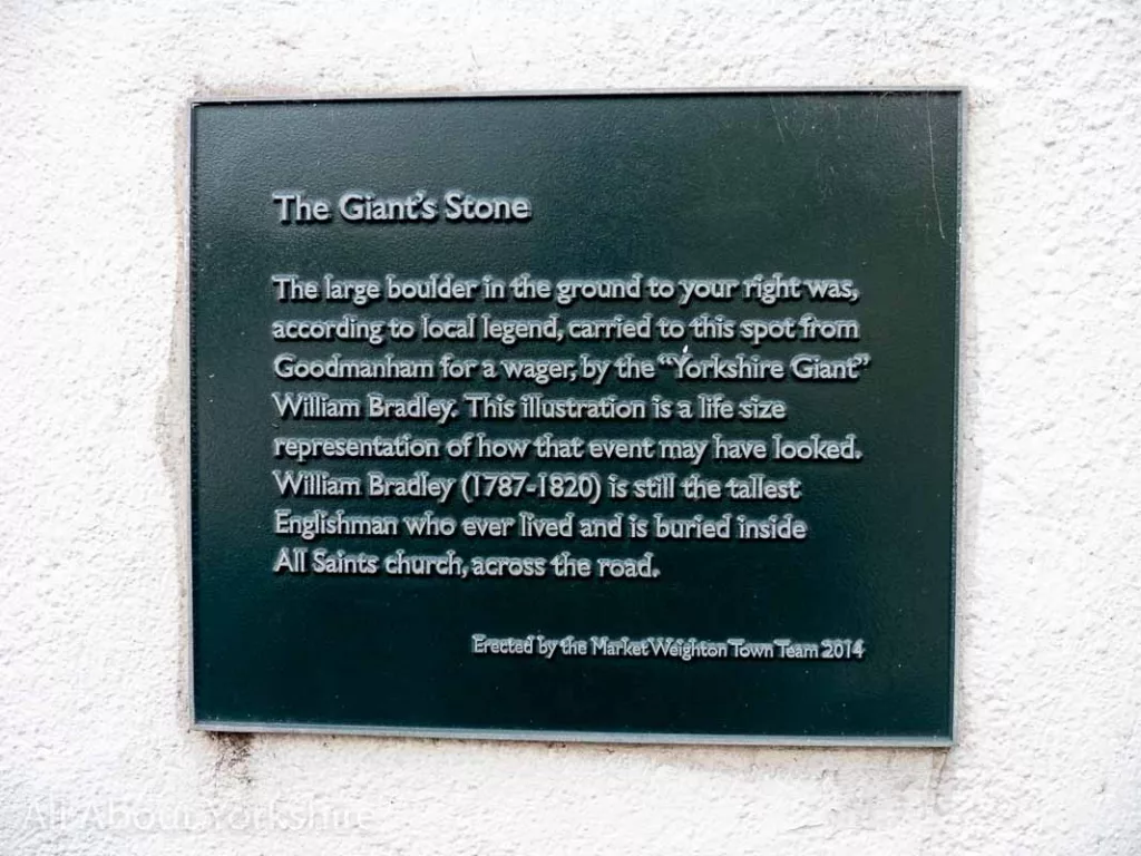 Plaque which reads: The Giant's Stone The large boulder in the ground to your right was, according to local legend, carried to this spot from Goodmanham for a wager, by the "Yorkshire Giant" William Bradley. This illustration is a life size representation of how that event may have looked. William Bradley (1787-1820) is still the tallest Englishman who ever lived and is buried inside All Saints church across the road. Erected by the Market Weighton Town team 2014