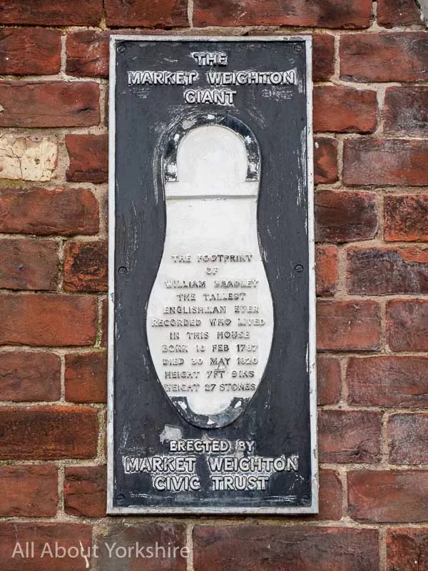 Black and white plaque with a life-size rendition of Willaim Bradley's shoe size.
Text reads:
"The Market Weighton Giant
The footrpint of William Bradley the tallest Englishman ever recorded who lived in this house born 11th Feb 1787 died 30th May 1820 Height 7FT 9INS Weight 27 stones"